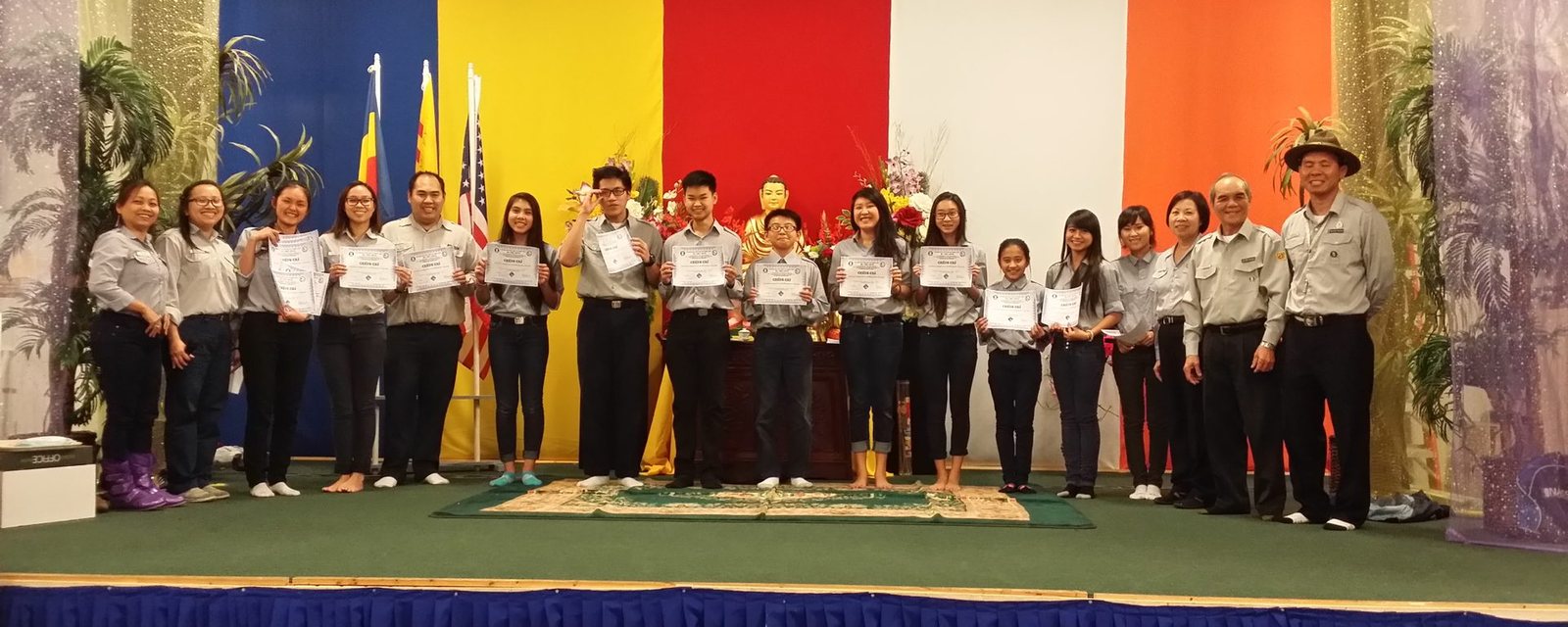 Hàng đoàn sinh GĐPT Huyền Quang đứng trên sân khấu cầm chứng chỉ, Sư Cô Tịnh Ngọc áo vàng đứng giữa, phía sau là cờ Phật giáo và cờ Hoa Kỳ