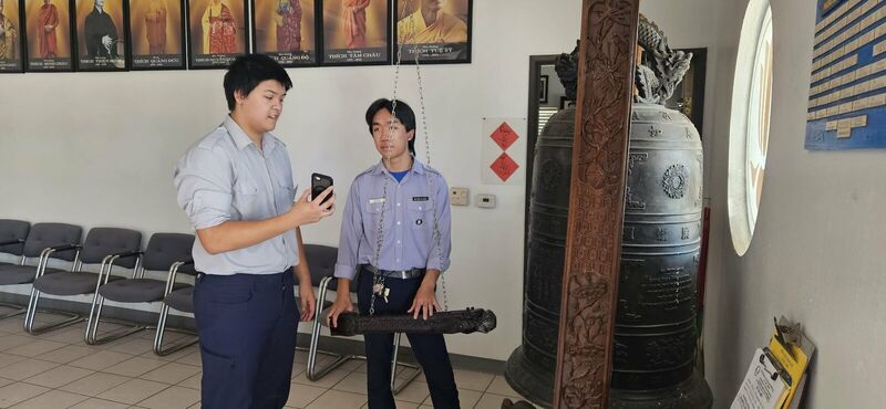 Hai em Huynh Trưởng tập sự bên đại hồng chung, một em dò lại nhịp trên điện thoại