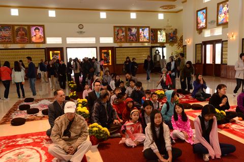 Các em ngồi trên thảm đỏ trong chánh điện, hoa cúc vàng, phụ huynh ngồi rìa