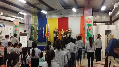 Nhìn từ sau lưng: các em đứng thành nhiều hàng hướng về bàn Phật, cờ Phật giáo trên tường