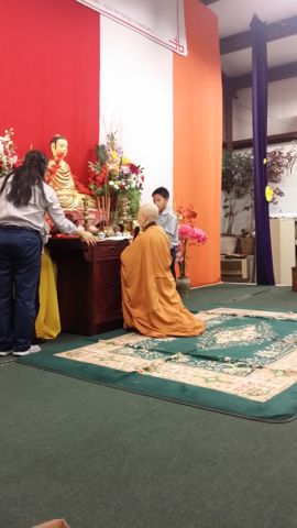 Sư Cô Tịnh Ngọc áo vàng quỳ trước bàn thờ Phật, một em đoàn sinh đứng bên bàn thờ, hoa và nhang đèn phía trên