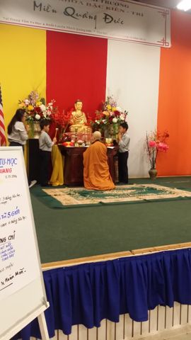 Nhìn từ xa: Sư Cô Tịnh Ngọc quỳ giữa thảm trước bàn Phật, hai em đứng hai bên, tấm bảng chương trình dựng bên trái sân khấu