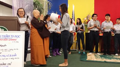 Tấm bảng trắng viết tay ghi Chương Trình Tu Học Huyền Quang 12.13.2014, cạnh bục đọc và Sư Cô Tịnh Ngọc áo vàng