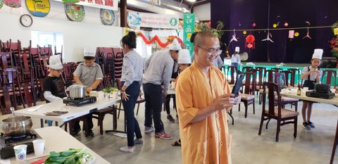 Một vị Thầy áo vàng cam đứng cười giữa hội trường, cầm điện thoại chụp các em đang nấu