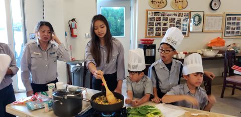 Chị Huynh Trưởng cầm đũa xào rau trên chảo, ba em Oanh Vũ đội mũ Chef Phú Quý đứng nhìn sát bên