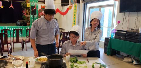 Đội Chef Quảng Bình: hai Huynh Trưởng đứng hai bên, em Ngành Thiếu ngồi giữa đọc công thức, chảo và rau trên bàn
