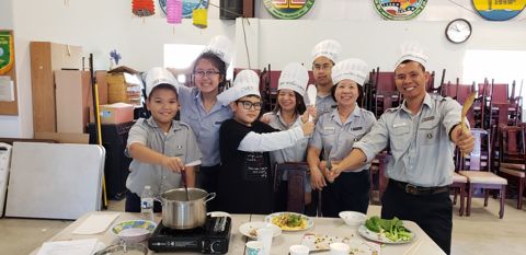 Một đội chụp chung, anh Huynh Trưởng giơ muỗng, các em cười, dĩa mì xào và dĩa rau trên bàn