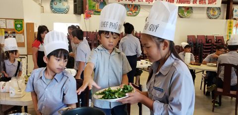 Một em Oanh Vũ bưng dĩa rau xanh đi ngang qua bàn, các em khác ngoái nhìn