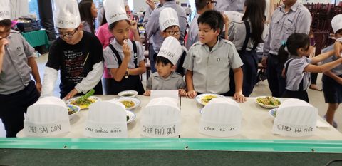 Các em nhỏ đứng sau bàn dài, năm chiếc mũ giấy Chef Sài Gòn, Quảng Bình, Phú Quý, Đà Lạt, Hà Nội xếp hàng, dĩa món sau mỗi mũ